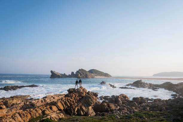 Mid distance image of couple standing on rocky coastline at beach. Male and female are enjoying the view of Plettenberg Bay. Scenic view of sea against clear sky.